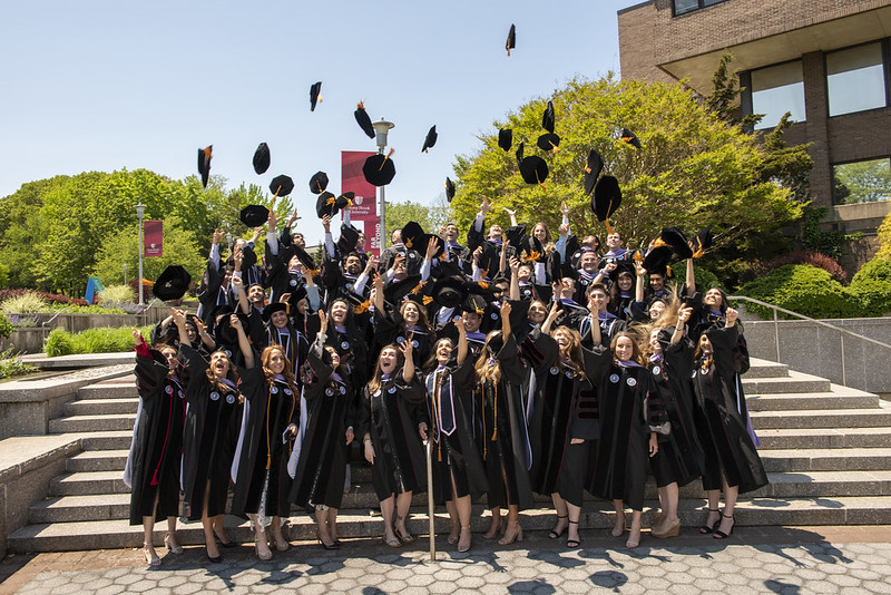 An outdoor group photo of Stony Brook School of Dental Medicine graduates in regalia tossing their graduation caps. They are standing on steps with trees in the background.